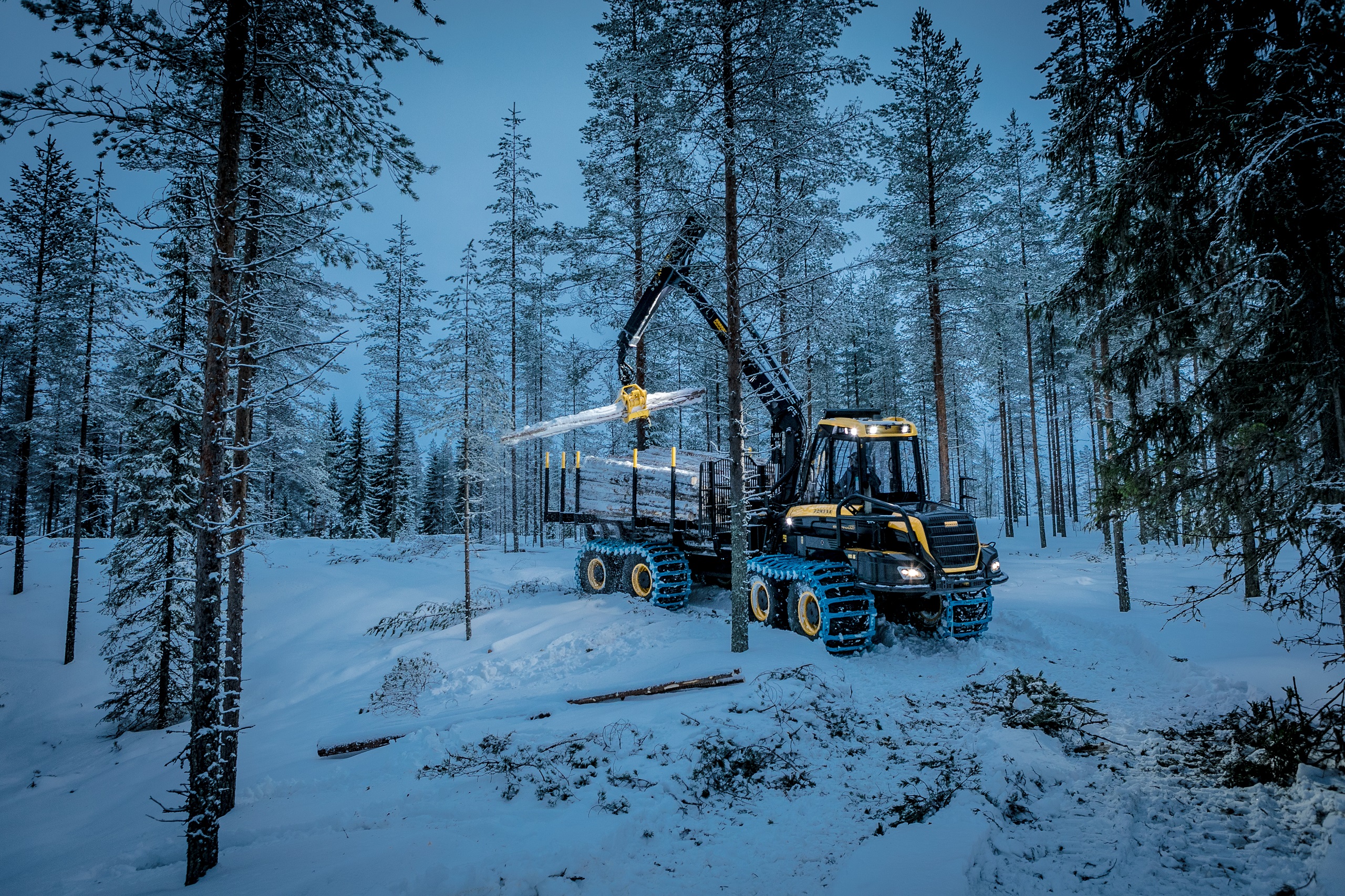 The picture depicts a snowy forest at night, with a yellow tractor in the centre loading cut trees onto its trailer.